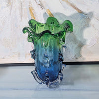 Glass vase with green top and blue bottom on a white surface with abstract painting in the background