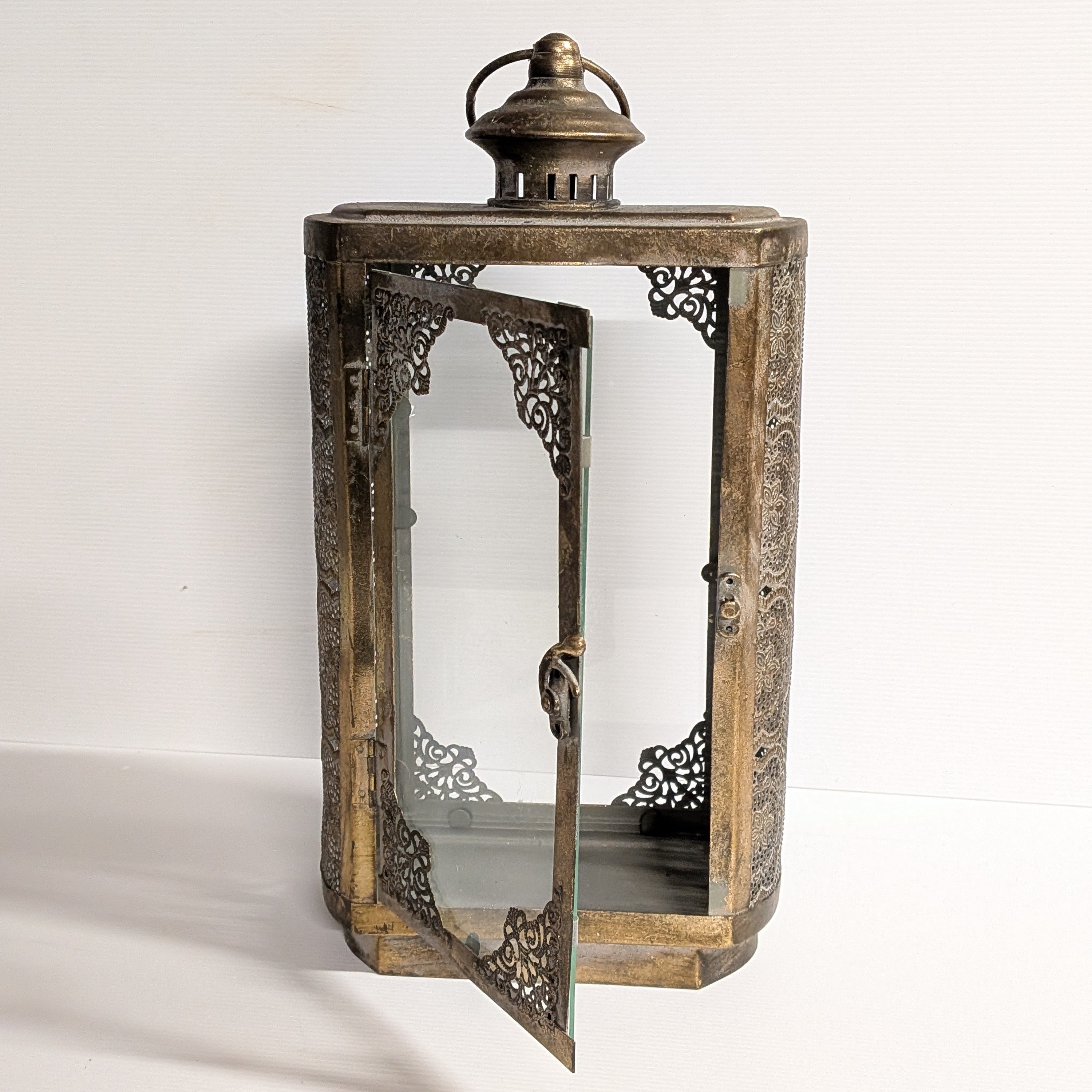 An antique-style lantern with a weathered bronze and gold-toned metal frame, featuring an open door with intricate filigree cutouts and a glass panel, set against a plain white background.