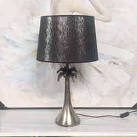 Table lamp with black shade and silver base on a white surface with abstract art in the background