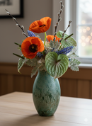 Green vase with orange and purple flowers arranged on a wooden table.